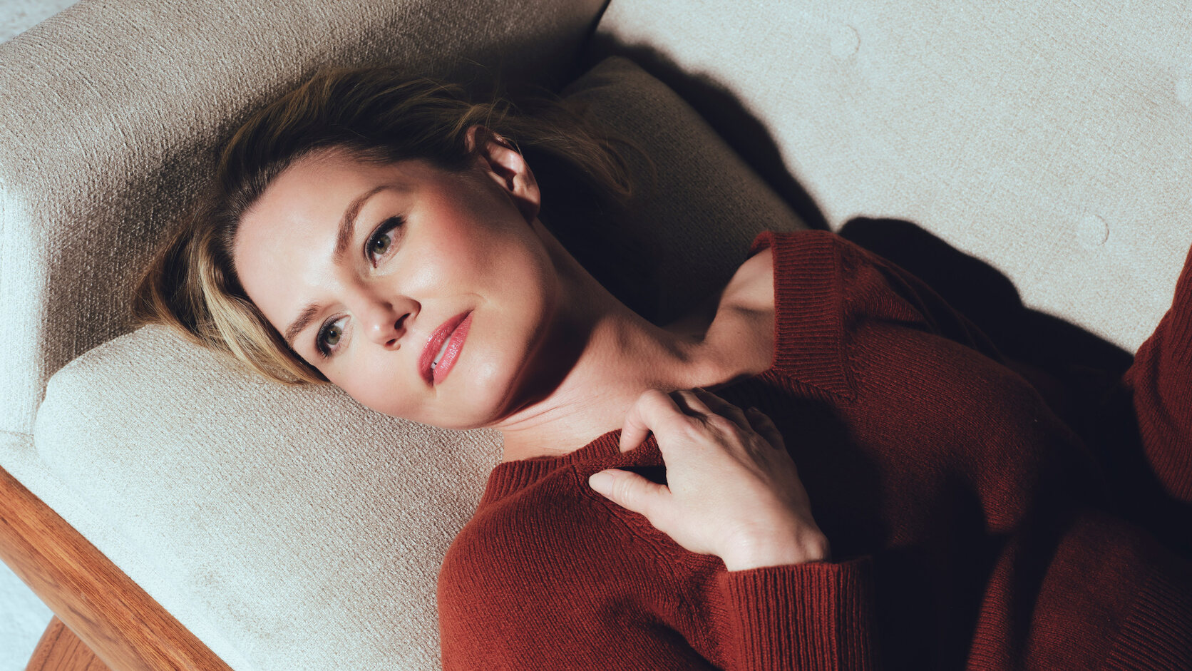 Jennifer Morrison lies back on a cream sofa with a wooden frame in a rust-red sweater, one hand resting at her collarbone, looking toward the camera.