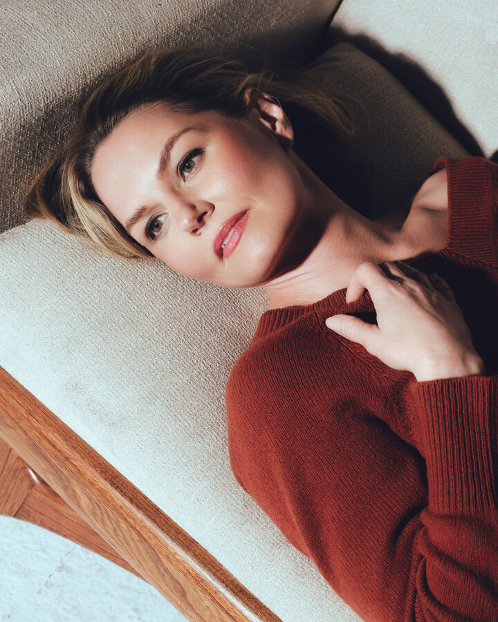 Jennifer Morrison lies back on a cream sofa with a wooden frame in a rust-red sweater, one hand resting at her collarbone, looking toward the camera.