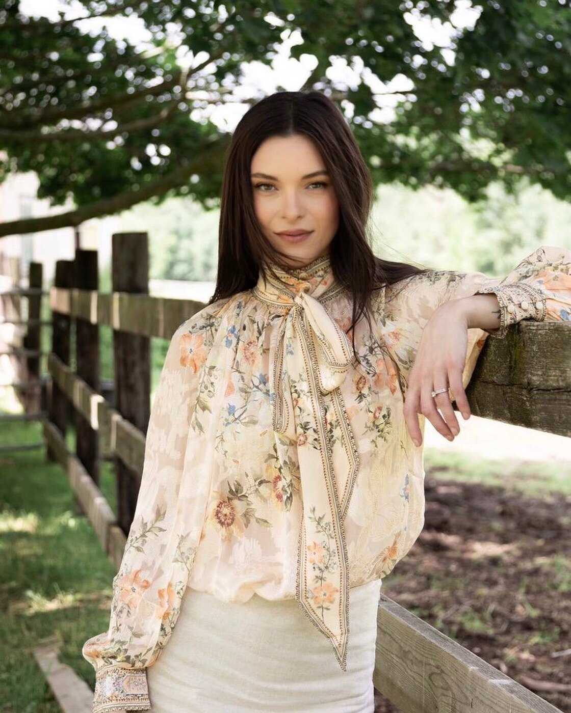 Jessica Parr, in a sheer cream floral blouse with a long tie-neck bow and a cream skirt, leans one arm on a weathered wooden fence beneath a shady tree and looks into the camera.