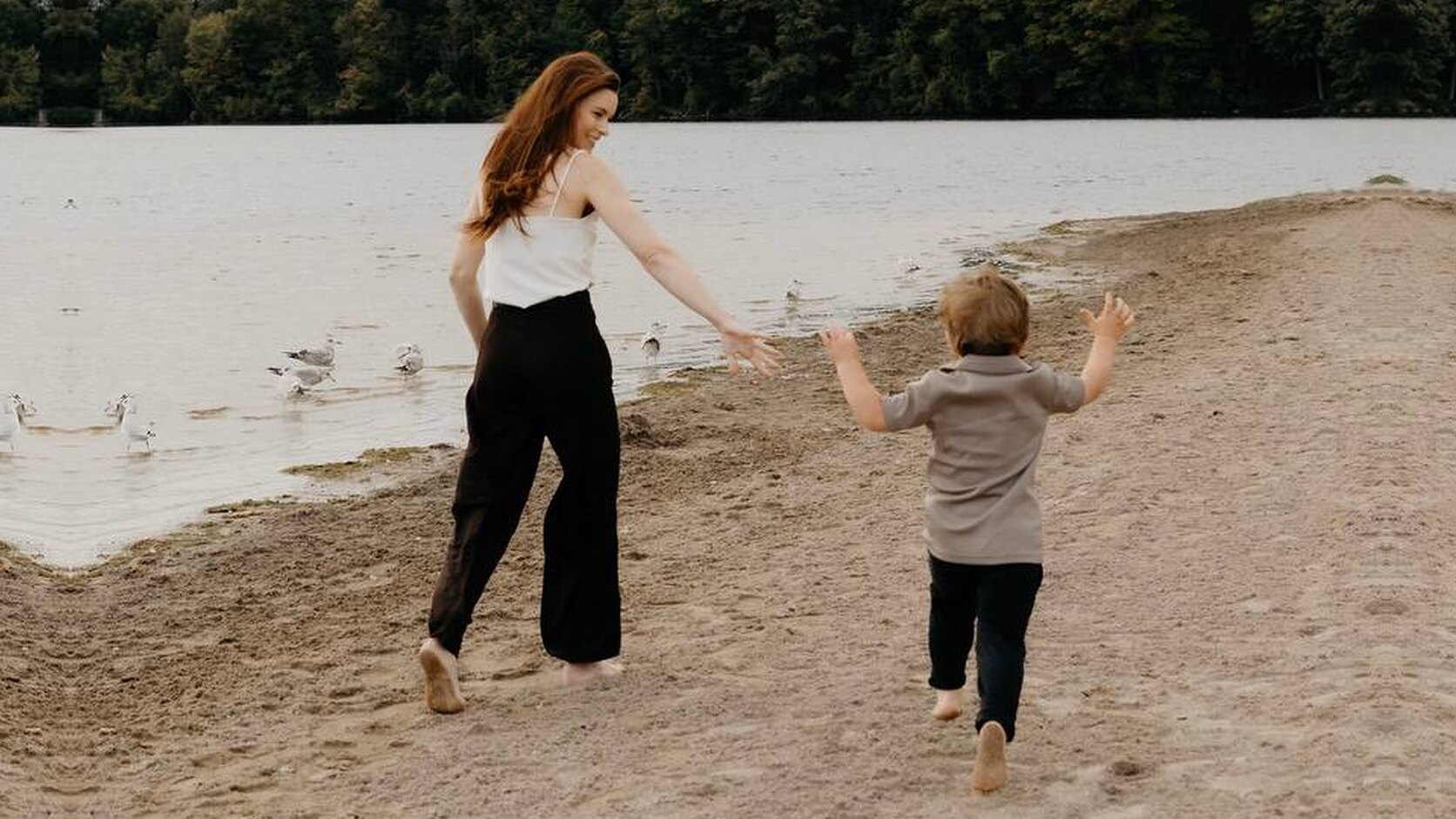 On a sandy lakeshore under an overcast sky, Jessica Parr, in a white camisole and wide black trousers, glances back with a smile and reaches out a hand as her young son runs barefoot after her, arms raised, past a flock of gulls at the water&rsquo;s edge.