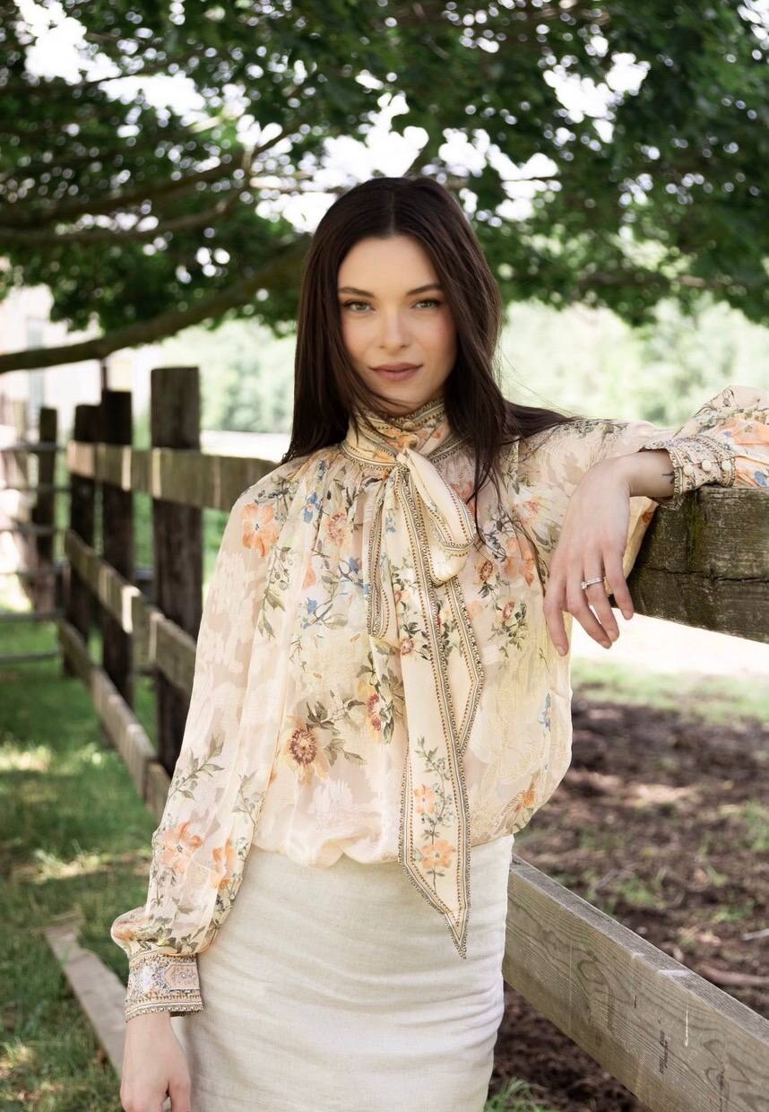 Jessica Parr, in a sheer cream floral blouse with a long tie-neck bow and a cream skirt, leans one arm on a weathered wooden fence beneath a shady tree and looks into the camera.