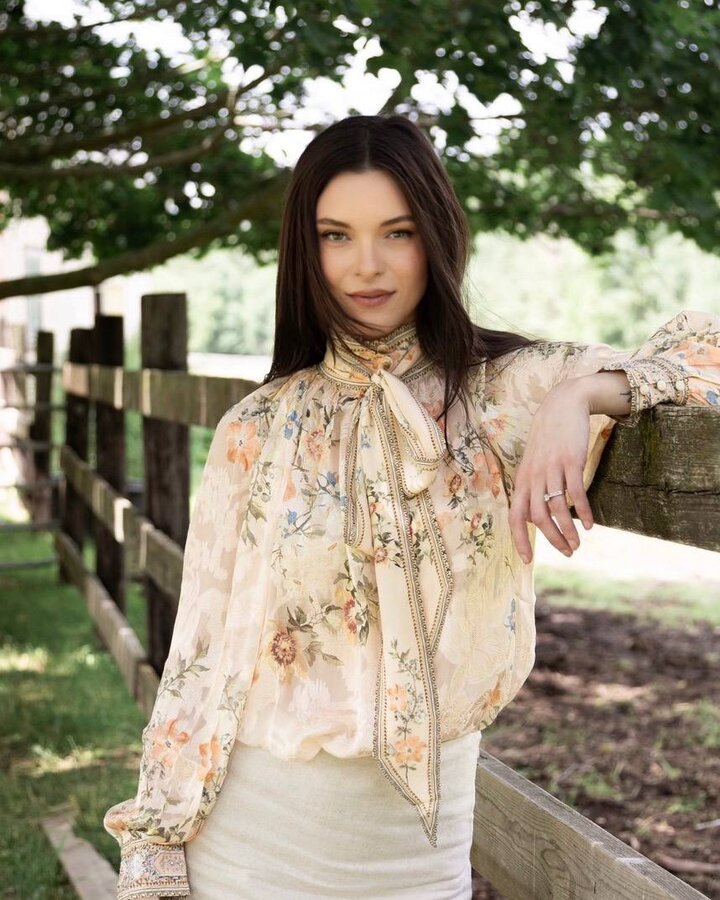 Jessica Parr, in a sheer cream floral blouse with a long tie-neck bow and a cream skirt, leans one arm on a weathered wooden fence beneath a shady tree and looks into the camera.