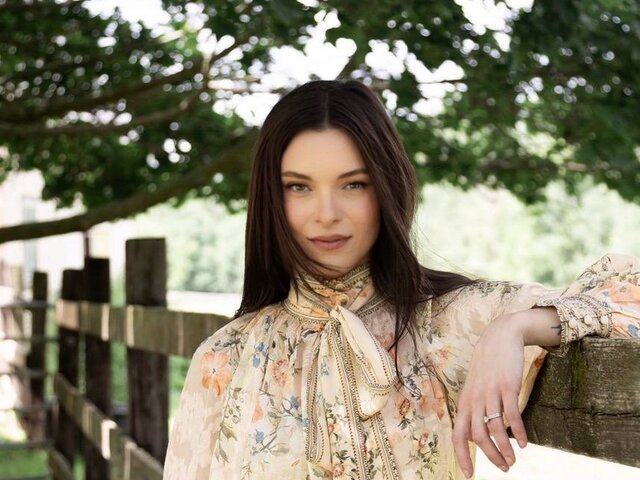 Jessica Parr, in a sheer cream floral blouse with a long tie-neck bow and a cream skirt, leans one arm on a weathered wooden fence beneath a shady tree and looks into the camera.