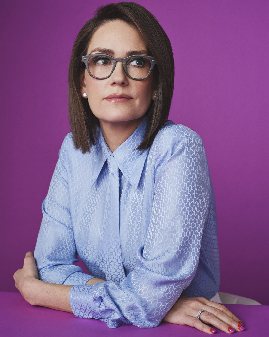 Jessica Tarlov in grey wood-frame glasses and a pale blue patterned blouse sits at a purple table against a matching purple backdrop, arms folded on the tabletop, glancing to the side.