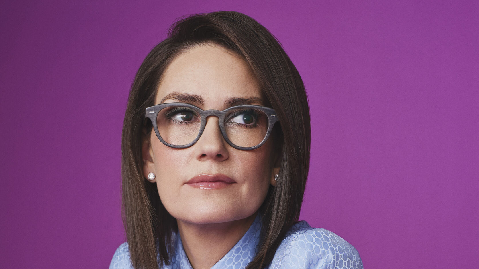 Jessica Tarlov in grey wood-frame glasses and a pale blue patterned blouse glances to the side against a bright purple backdrop.