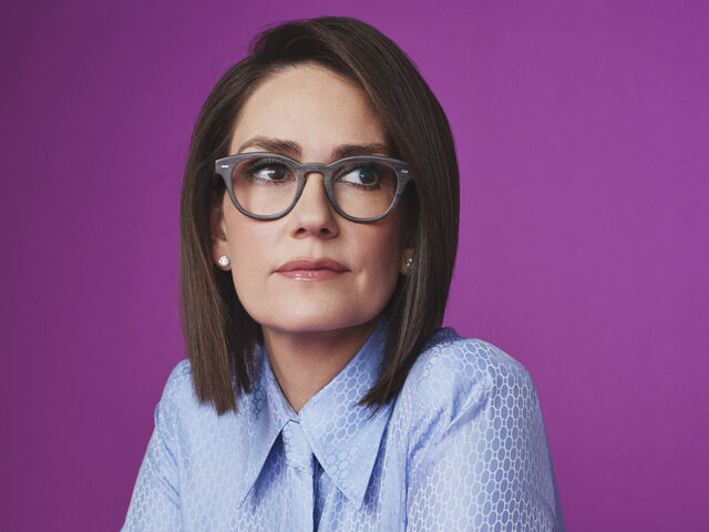 Jessica Tarlov in grey wood-frame glasses and a pale blue patterned blouse sits at a purple table against a matching purple backdrop, arms folded on the tabletop, glancing to the side.