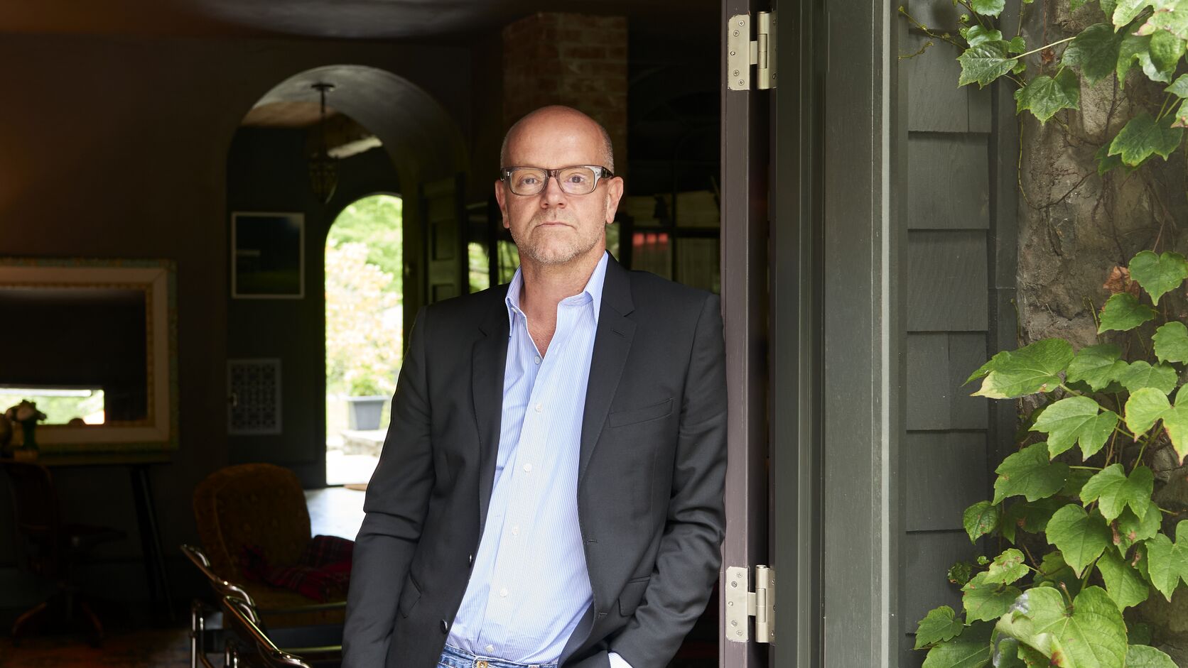 Jonathan Van Meter, in glasses, a dark blazer, light blue shirt and jeans, leans in an open doorway framed by ivy, a dim wood-floored room with arched doorways behind him.