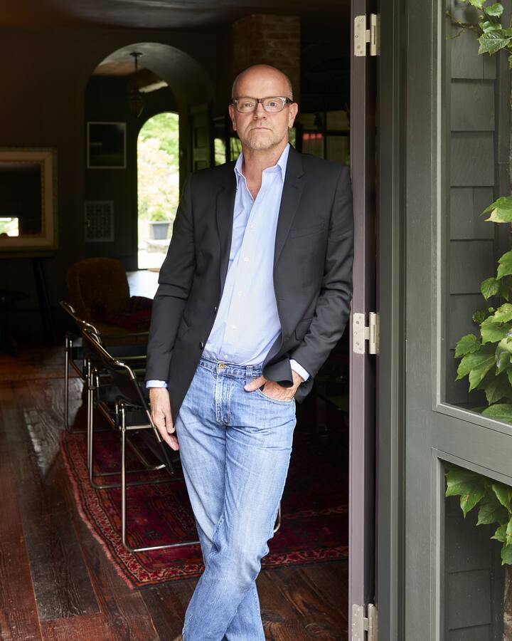 Jonathan Van Meter, in glasses, a dark blazer, light blue shirt and jeans, leans in an open doorway framed by ivy, a dim wood-floored room with arched doorways behind him.