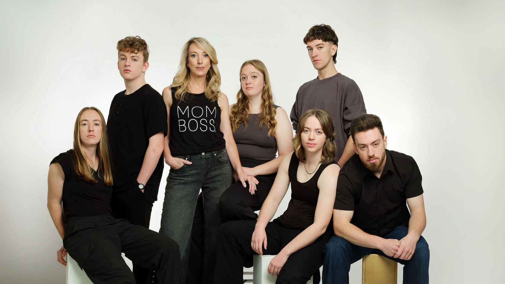 Julie Cole stands in a black MOM BOSS tank top surrounded by her six grown children, all in black and grey against a white studio backdrop.