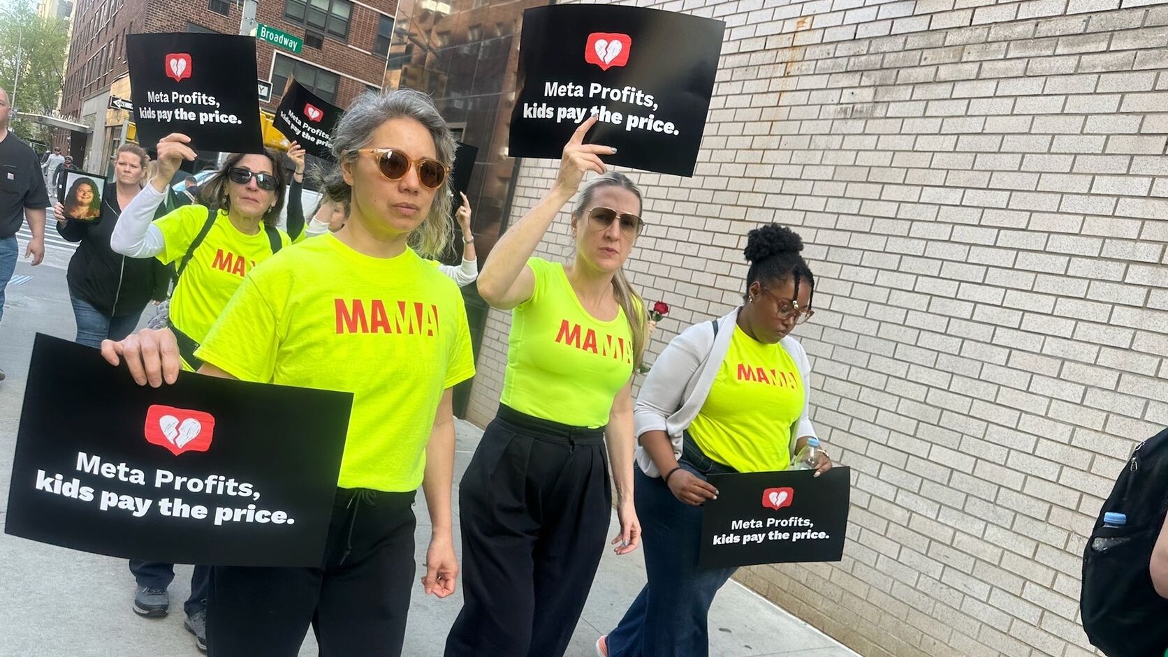 Mothers in neon yellow MAMA t-shirts march down a city sidewalk holding black signs that read "Meta Profits, kids pay the price."
