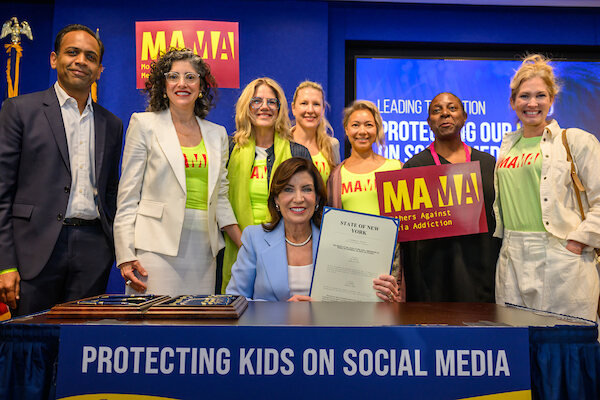 New York Governor Kathy Hochul, seated at a desk, holds up the signed state bill as MAMA members in yellow shirts stand behind her beneath a banner reading Protecting Kids on Social Media.