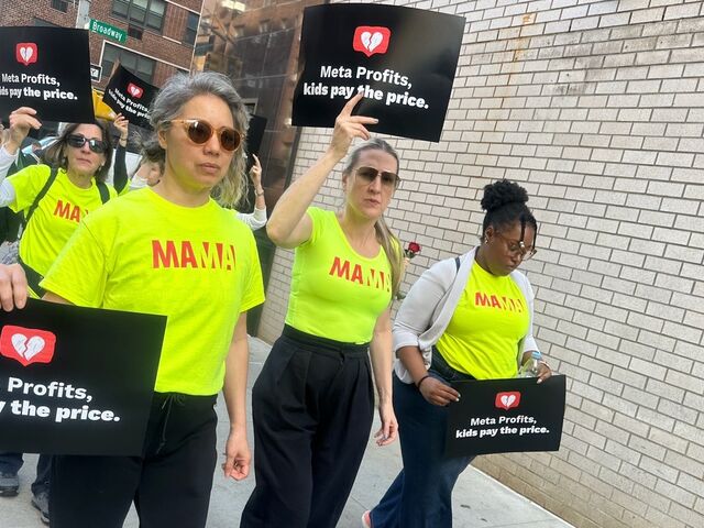 Mothers in neon yellow MAMA t-shirts march down a city sidewalk holding black signs.