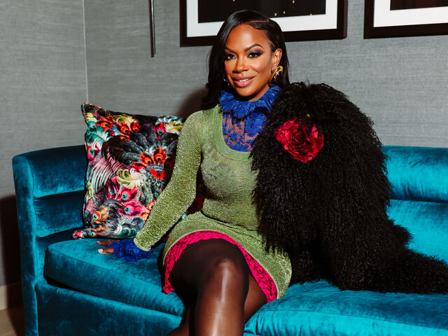 Kandi Burruss kneels on a teal velvet sofa in a green metallic mini dress with blue lace cuffs and collar, beneath the Momé September 2026 cover lines.