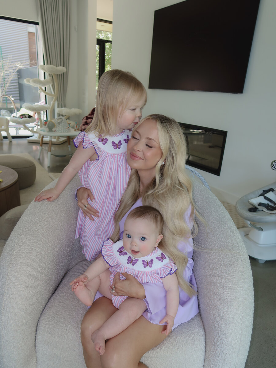 Kay Dudley sits in a cream armchair holding her baby daughter on her lap while her toddler leans in to kiss her cheek, both girls in matching pink butterfly dresses.