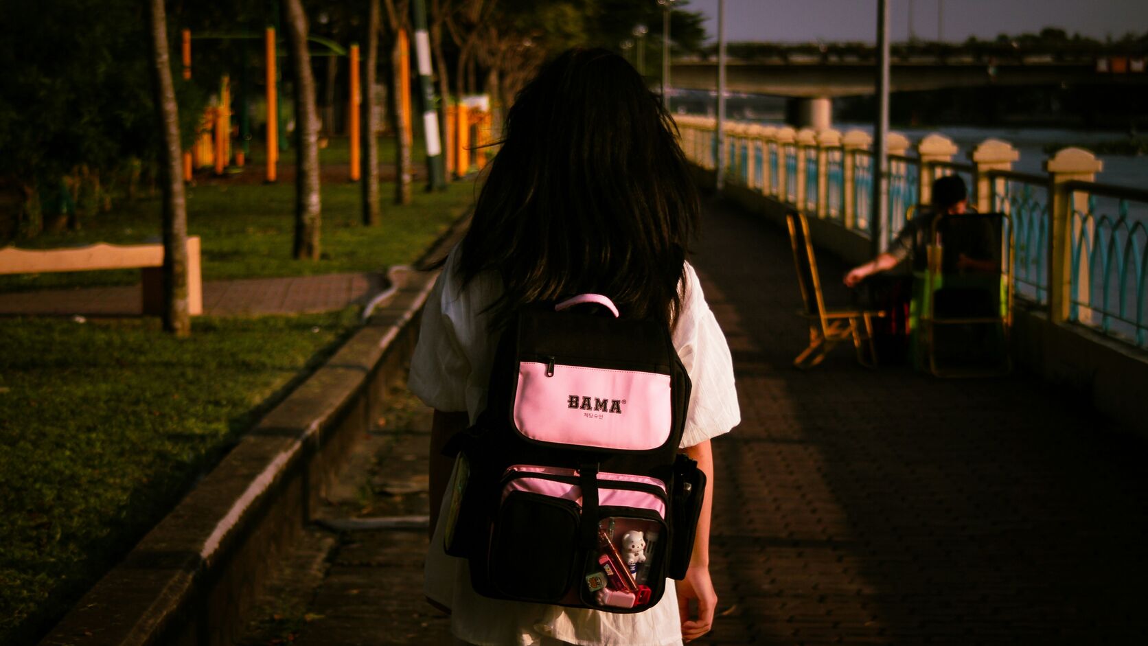A girl in a white dress walks away down a riverside path at golden hour, a pink and black school backpack on her shoulders.