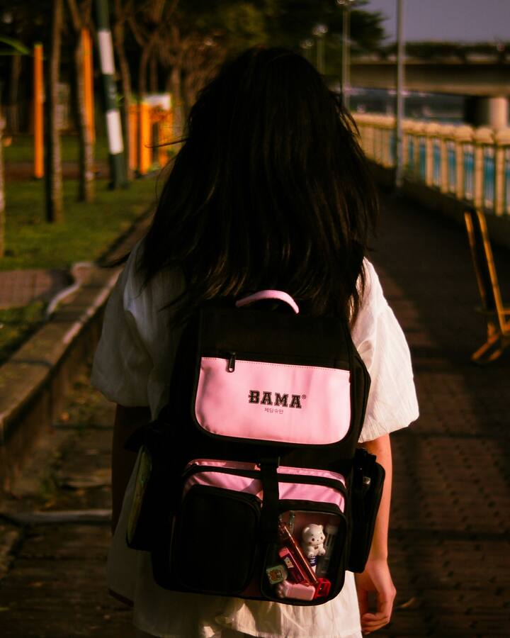 A girl in a white dress walks away down a riverside path at golden hour, a pink and black school backpack on her shoulders.