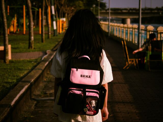 A girl in a white dress walks away down a riverside path at golden hour, a pink and black school backpack on her shoulders.