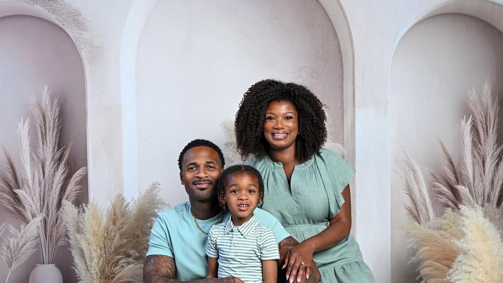 LaShonda Unseld Hopkins, her husband, and their son Major sit together in matching sage green against a white arched backdrop with pampas grass.