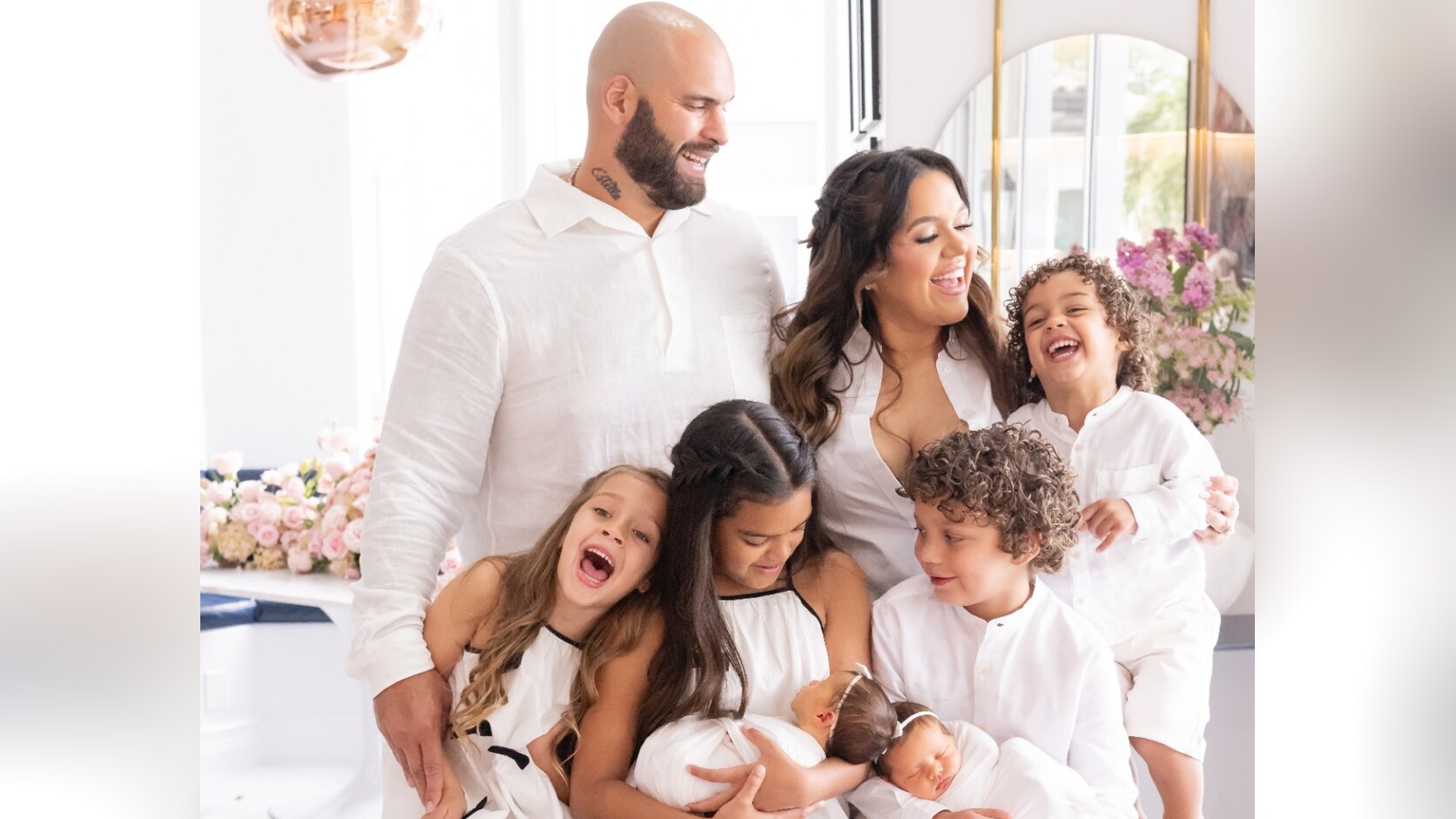 Lawrence and Andrea Guy stand with their six children, all dressed in white, in a bright living room, the newborn twins held by their older siblings.