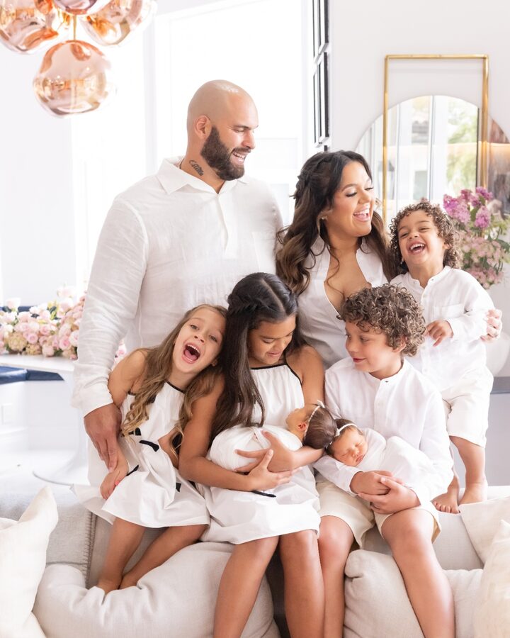 Lawrence and Andrea Guy stand with their six children, all dressed in white, in a bright living room, the newborn twins held by their older siblings.