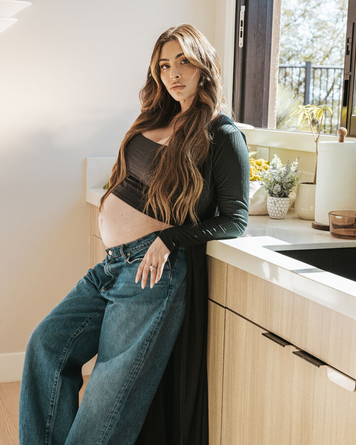 Leila Hormozi, pregnant, in a black long-sleeve crop top and wide-leg jeans with long, wavy hair, leans against a sunlit kitchen counter and looks into the camera.