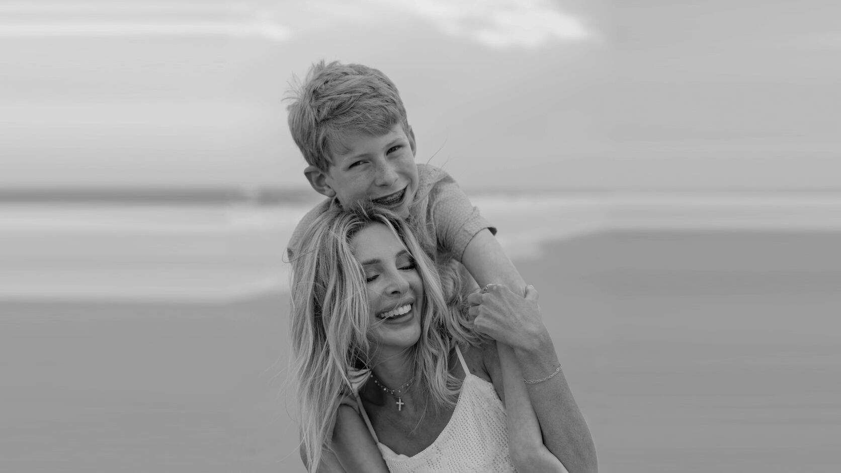 In black and white on a beach, Lindsie Chrisley laughs with her eyes closed in a white crochet sundress as her son hugs her from behind, his arms around her shoulders, grinning at the camera.