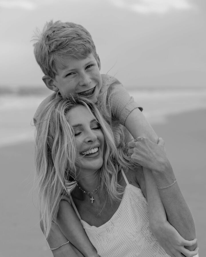 In black and white on a beach, Lindsie Chrisley laughs with her eyes closed in a white crochet sundress as her son hugs her from behind, his arms around her shoulders, grinning at the camera.
