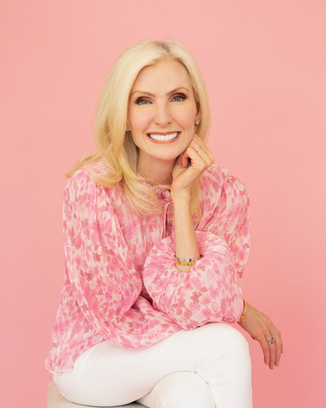 Lora McLaughlin Peterson sits against a pink backdrop in a pink floral blouse and white jeans, chin resting on one hand, smiling at the camera.