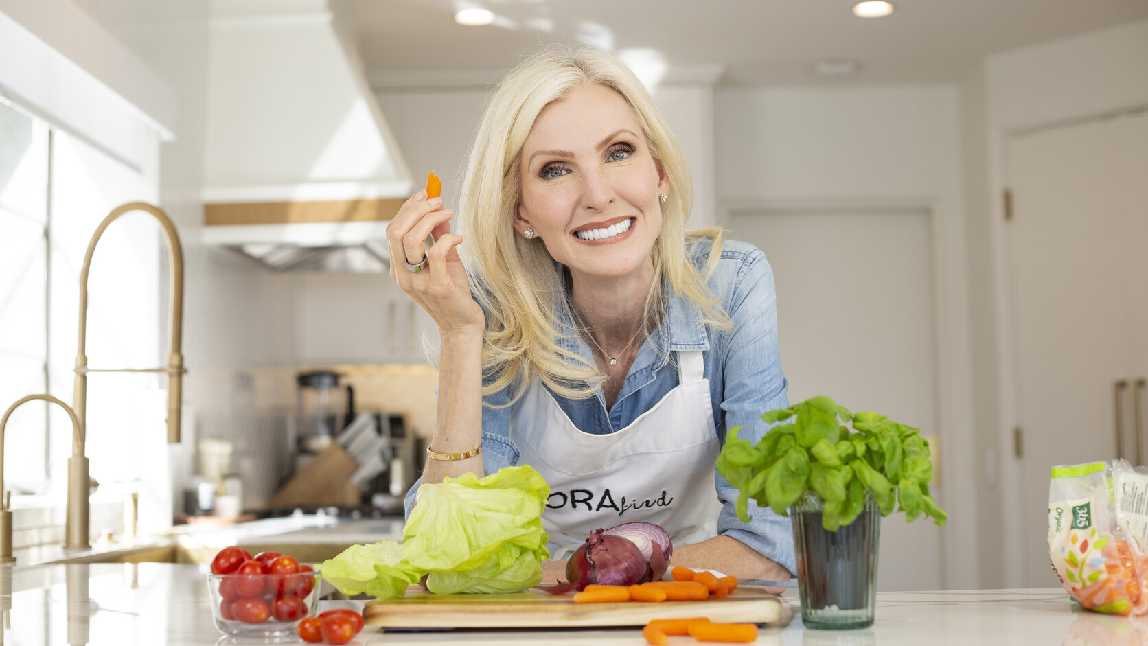 Lora McLaughlin Peterson leans on a white kitchen island in a chambray shirt and a LORAfied apron, holding up a carrot stick and smiling, with tomatoes, lettuce, a red onion and a pot of basil in front of her.