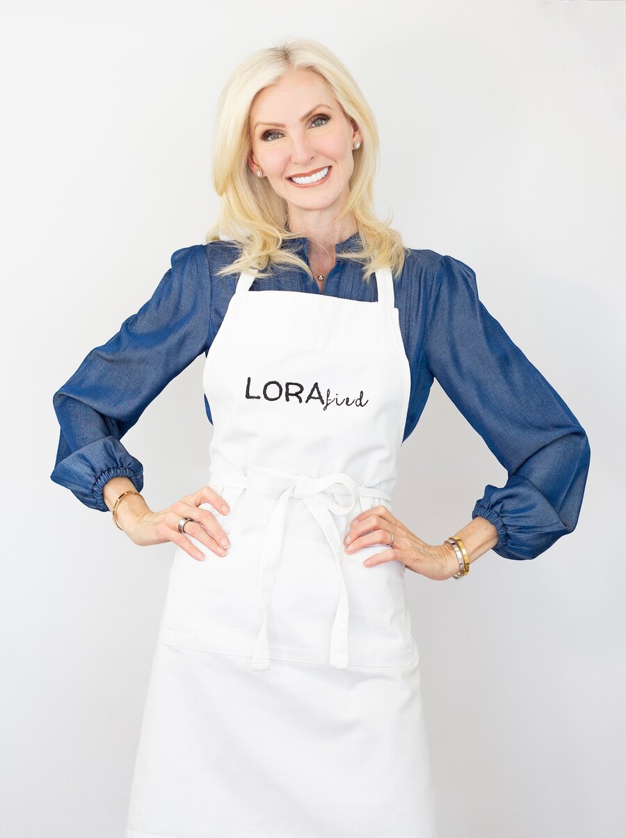 Lora McLaughlin Peterson stands against a white backdrop in a denim blouse and a white LORAfied apron, hands on her hips, smiling at the camera.