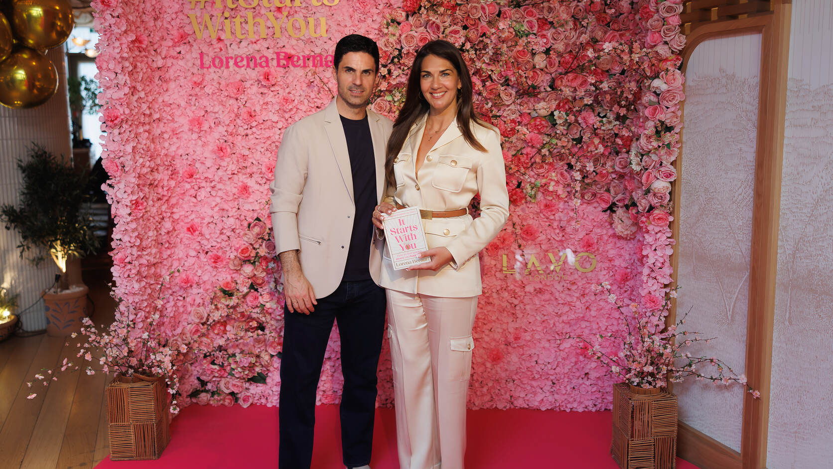 Lorena Bernal in a cream suit holds a copy of her book It Starts With You in front of a pink flower wall at her London book launch, her husband standing beside her.