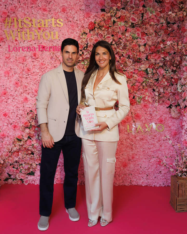 Lorena Bernal in a cream suit holds a copy of her book It Starts With You in front of a pink flower wall at her London book launch.