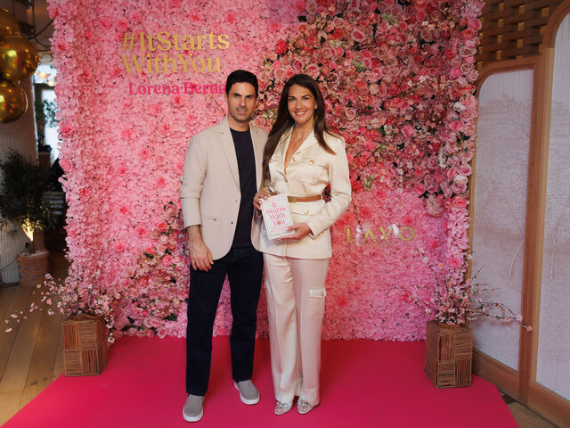 Lorena Bernal in a cream suit holds a copy of her book It Starts With You in front of a pink flower wall at her London book launch.