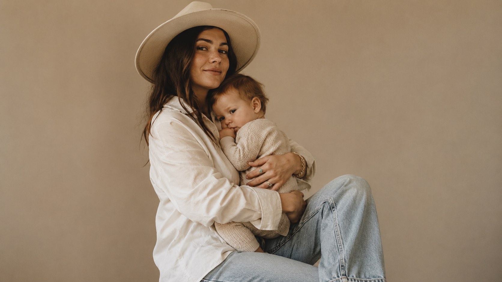 A mother in a wide-brimmed hat and white shirt holding her baby close against a warm neutral backdrop.