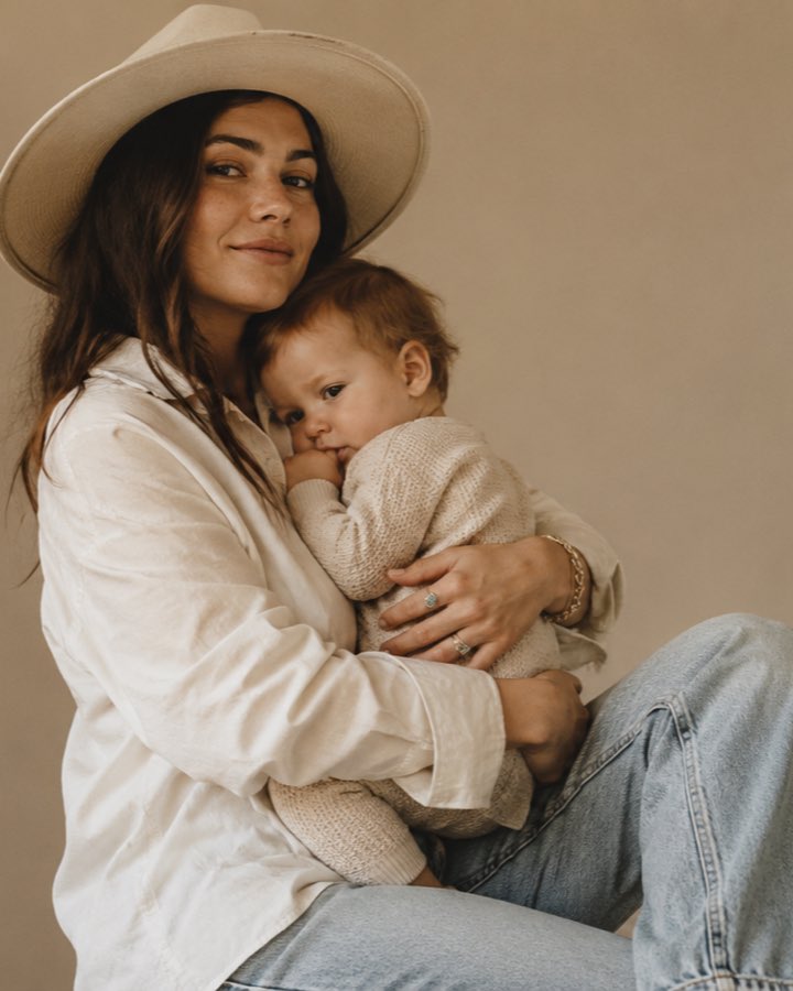 A mother in a wide-brimmed hat and white shirt holding her baby close against a warm neutral backdrop, both calm in soft studio light.
