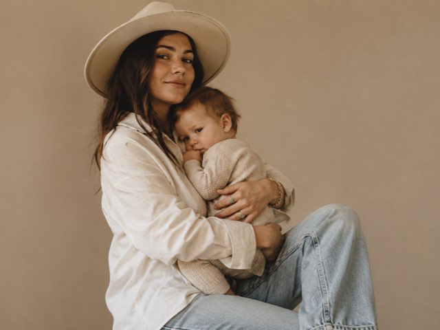 A mother in a wide-brimmed hat holds her baby in soft studio light.