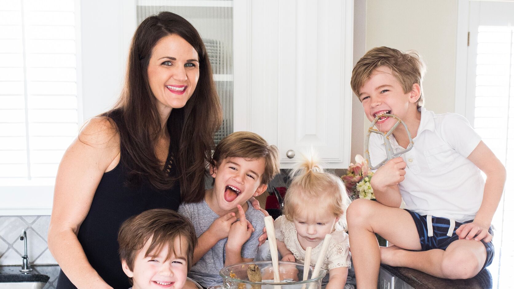 Maegan Brown stands in her white kitchen with her four children around a mixing bowl of cookie dough, one son on the counter licking the paddle attachment.