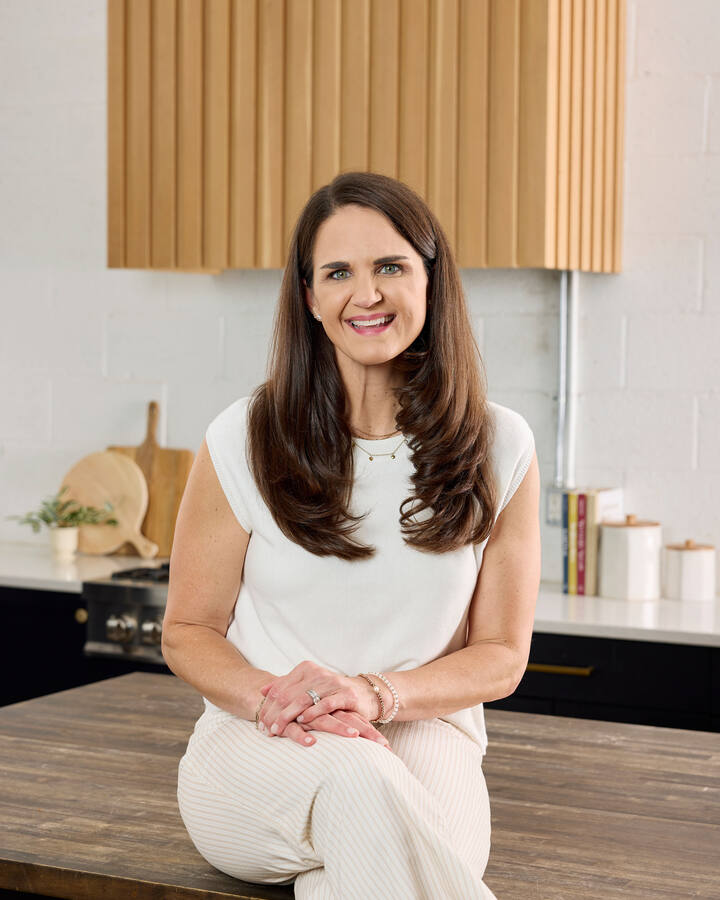 Maegan Brown sits on a wooden kitchen island in a cream sleeveless knit top and striped trousers, hands clasped, smiling at the camera.