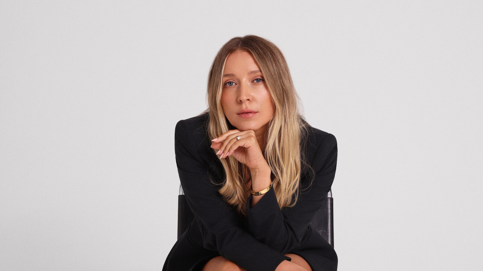 Margarita Nazarenko sits on a chrome cantilever chair against a pale grey studio backdrop in an oversized black blazer, her chin resting on her hand.