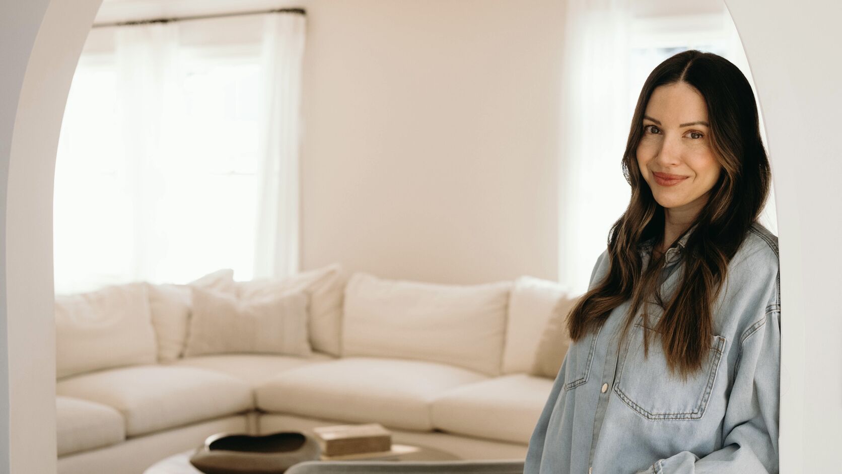 Maria Roberts stands in a light-wash denim shirt and jeans beneath a white archway, a cream sectional and sunlit living room behind her.