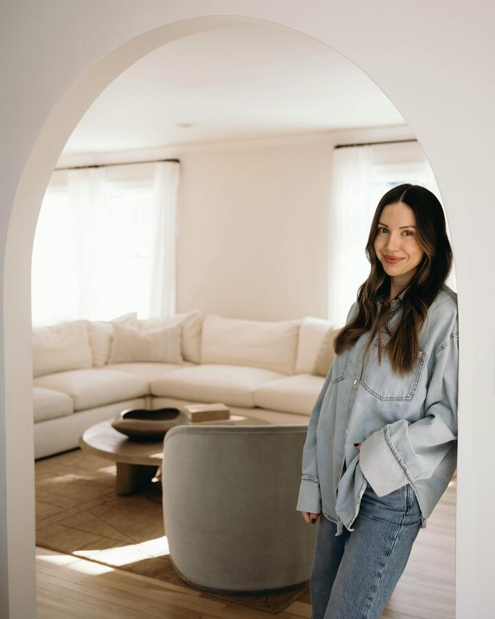 Maria Roberts stands in a light-wash denim shirt and jeans beneath a white archway, a cream sectional and sunlit living room behind her.