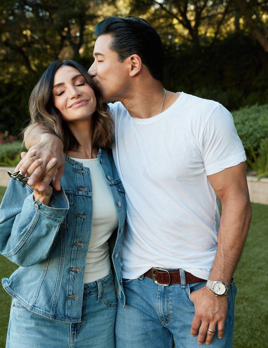 Mario Lopez, in a white T-shirt and jeans, kisses Courtney Lopez on the temple as she smiles with her eyes closed in a denim jacket, in a sunlit garden.