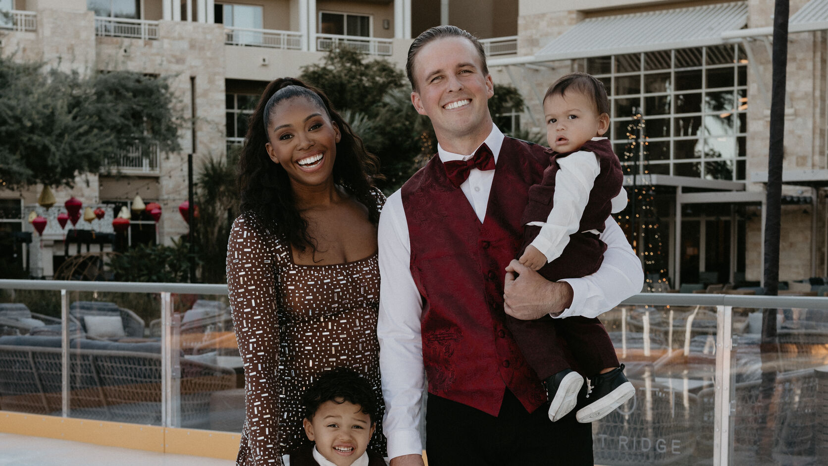 Mariyah Gerber in a brown patterned skating dress and her husband in a burgundy vest and bow tie smile on an outdoor ice rink, he holding their baby son on his hip and their older son standing in front of them in a matching burgundy vest.