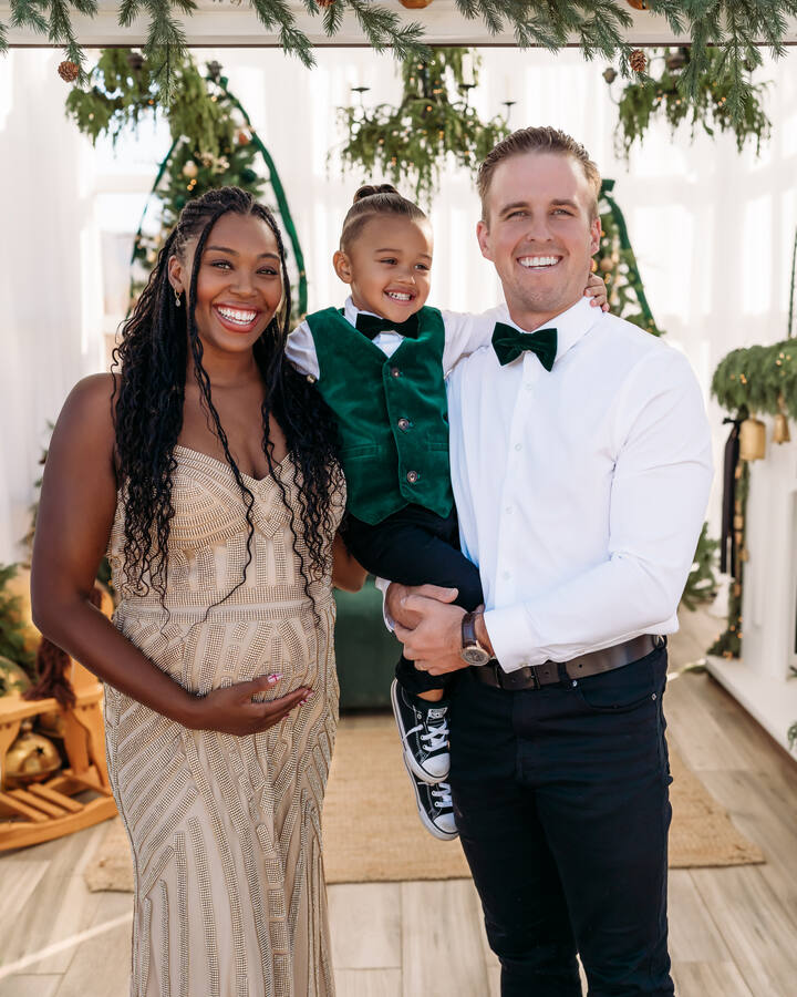 Mariyah Gerber, pregnant in a beaded champagne gown, smiles beside her husband, who holds their young son in a green velvet vest and bow tie beneath a pine garland.