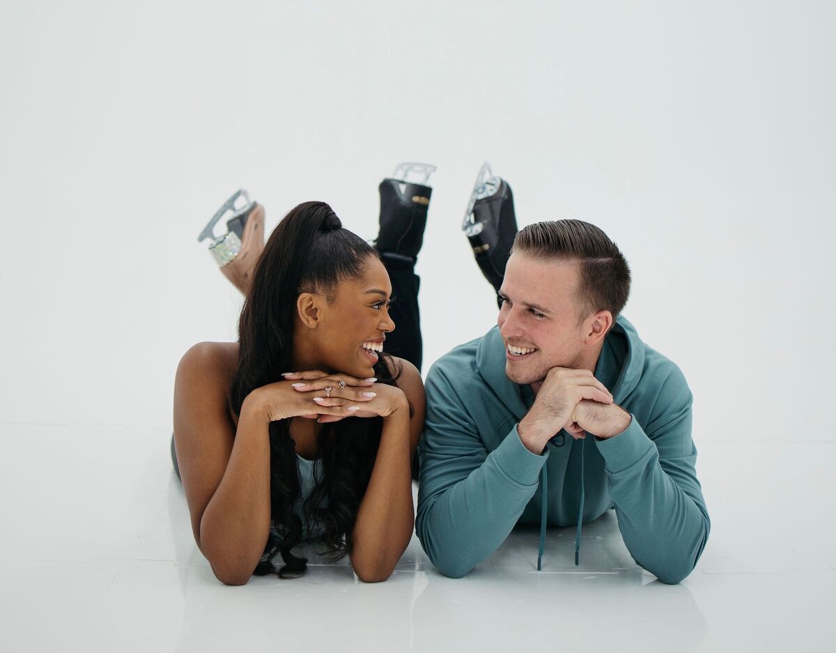 Mariyah Gerber and her husband lie on their stomachs on white ice in their skates, chins resting on their clasped hands, grinning at each other.