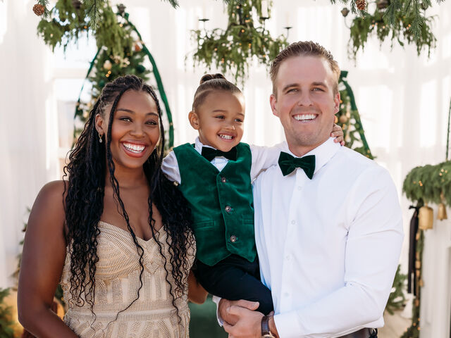 Mariyah Gerber in a brown patterned skating dress and her husband in a burgundy vest and bow tie smile on an outdoor ice rink, he holding their baby son on his hip and their older son standing in front of them in a matching burgundy vest.