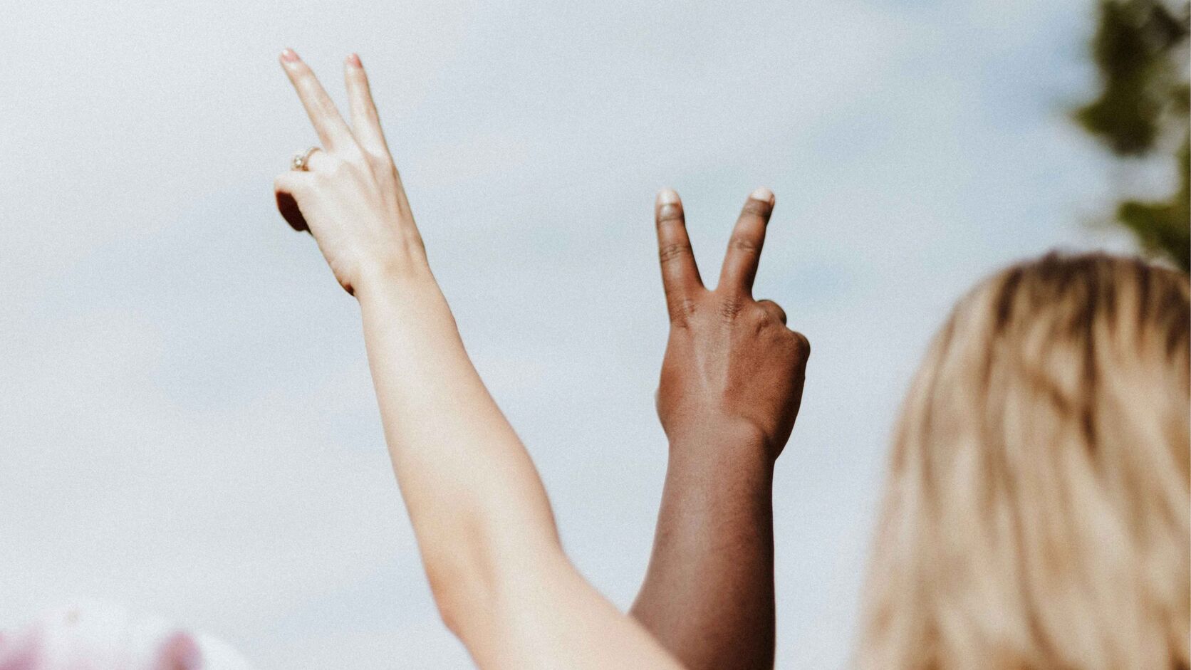 Two raised arms of different skin tones make peace signs against a pale blue sky, beside the back of a blonde woman's head.