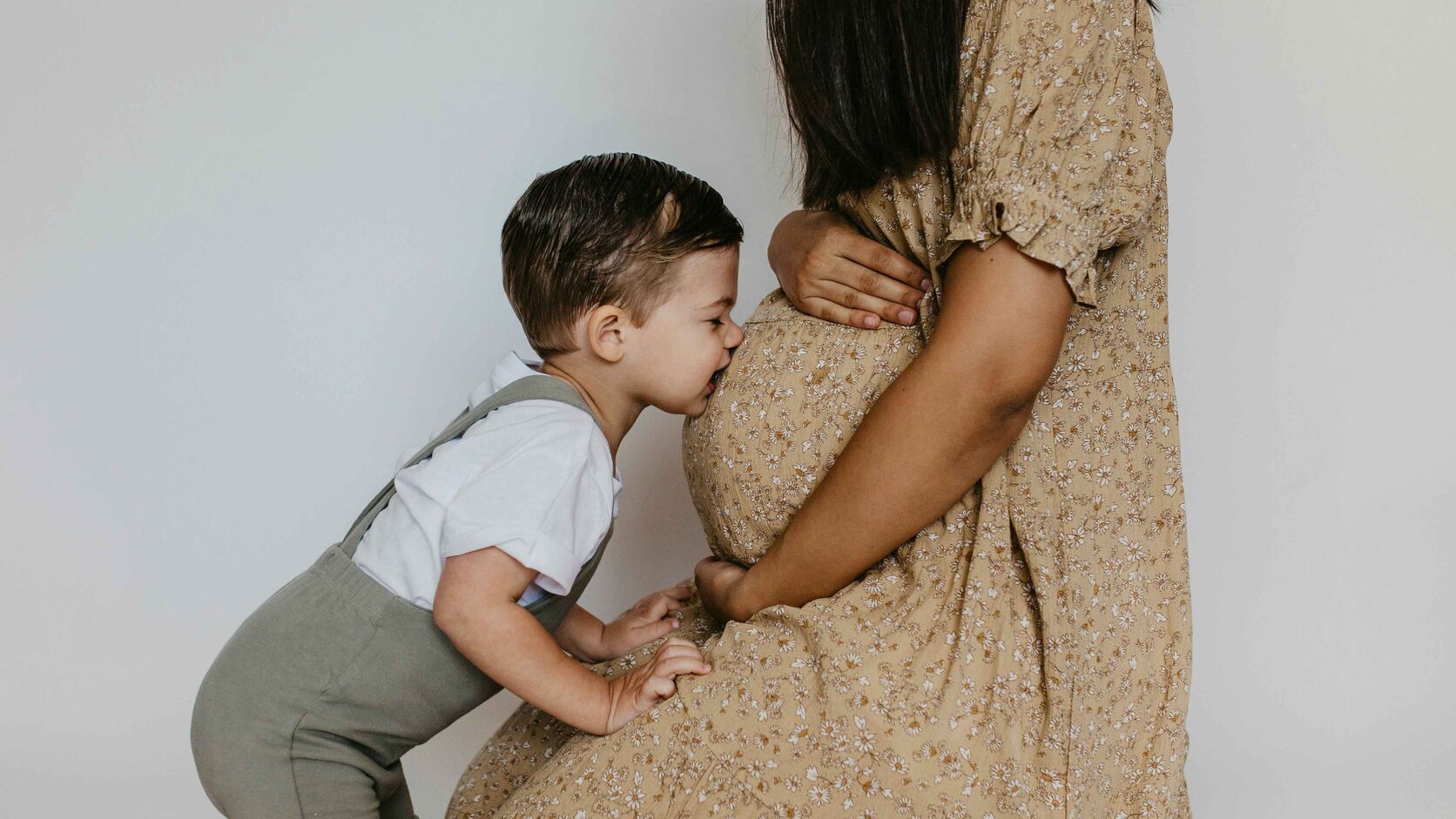 A smiling toddler in a white shirt and suspenders kisses his pregnant mother's belly, her hand resting gently on the bump.