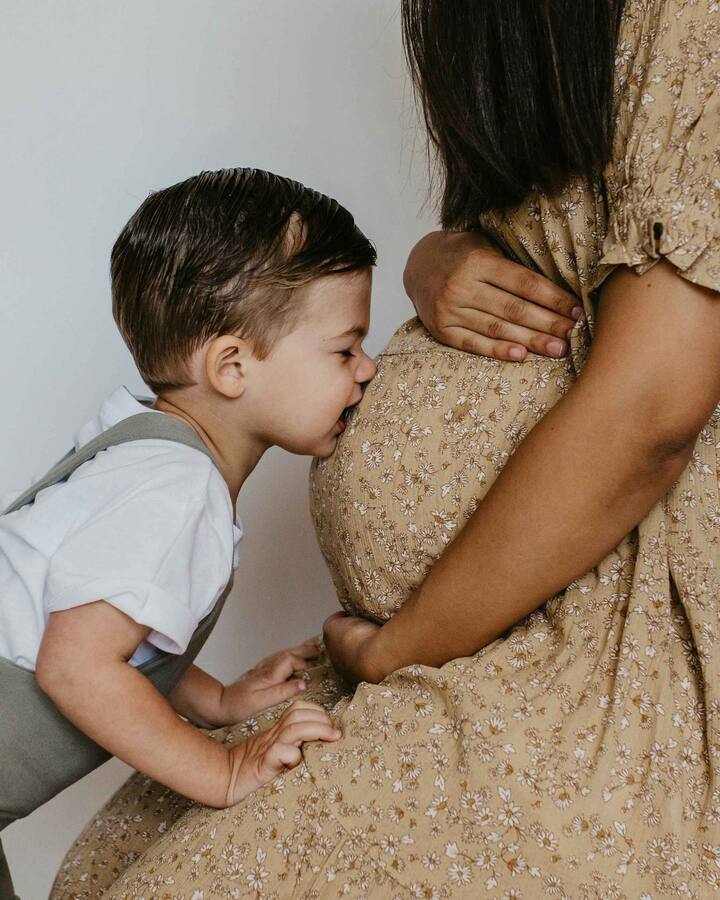 A smiling toddler in a white shirt and suspenders kisses his pregnant mother's belly, her hand resting gently on the bump.
