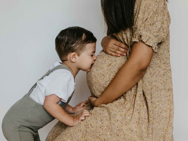 A smiling toddler in a white shirt and suspenders kisses his pregnant mother's belly, her hand resting gently on the bump.